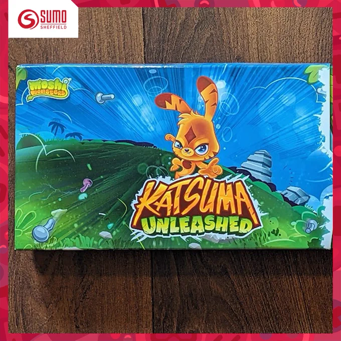 On a wooden table is the packaging for 'Moshi Monsters: Katsuma Unleashed.' The packaging shows a grassy environment with a blue sky. In the centre is the character 'Katsuma' a cute looking orange rabbit like creature from the Moshi Monsters franchise.