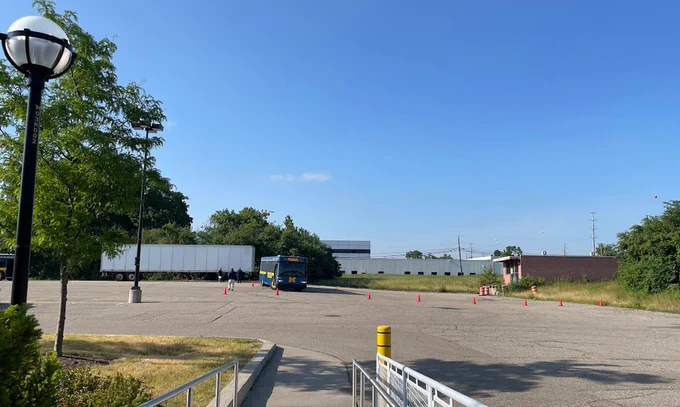 Open parking lot view from a loading dock ramp of a blue Michigan bus training with orange cones on the ground. Another bus is situated to the left of frame.