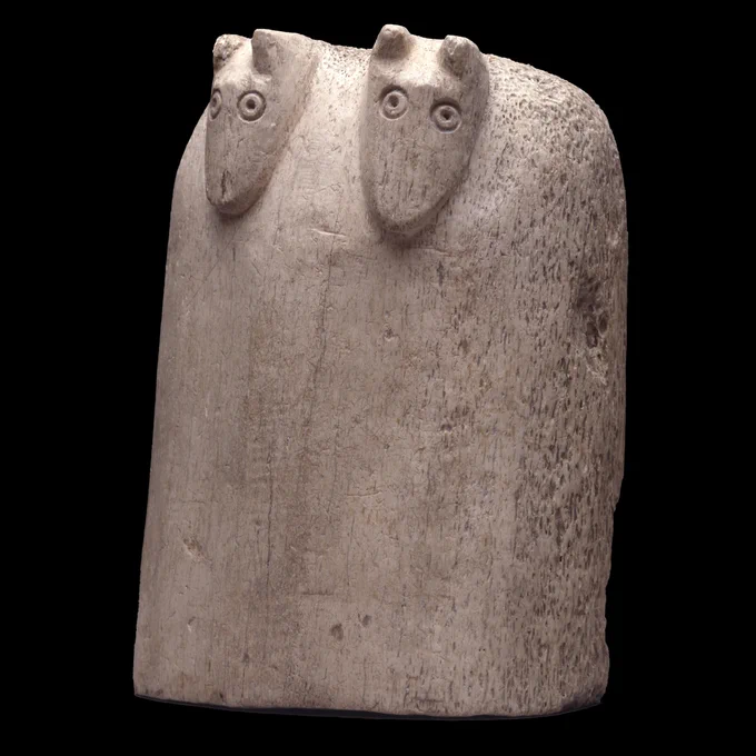A whalebone chess piece carved almost like a smoothed square with a rounded top. It is decorated with the simple heads of two small animals with ring-and-dot eyes, as if to look like two creatures were stood close together.