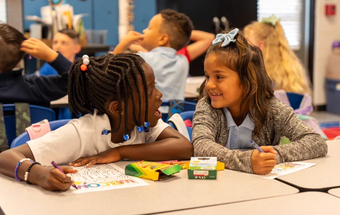 Two students sit at desks and talk to one another.