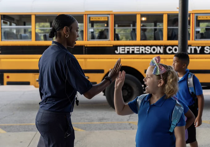 An adult and a student do a high five with a bus in the background.