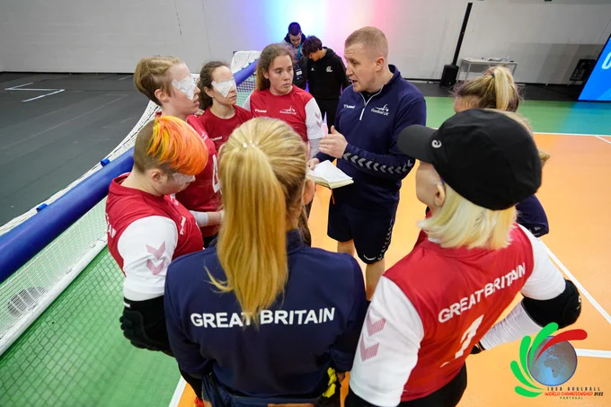 A group of GB Women players and their coaches stand at the goal end for a team talk during a game. Meme is on the left of the image.