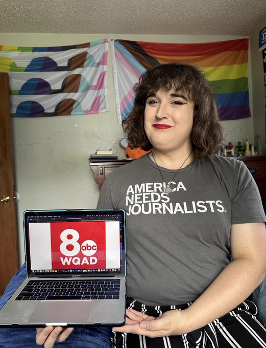 Nora, sitting in her bedroom, holding a laptop displaying the logo of WQAD 8 news. She’s wearing a gray tee shirt that reads “America Needs Journalists” in white font, with the shirt tucked into her black-and-white striped pants.