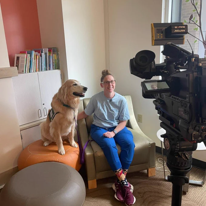 One of our facility dogs sits with an adult in front of a news camera. 