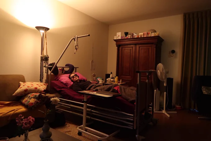 On the picture you see a person lying in a bed in a living room. The person in wearing sunglasses and headphones. You can see a cubbert behind the bed, a couch next to the bed and a fan. The curtains are closed.