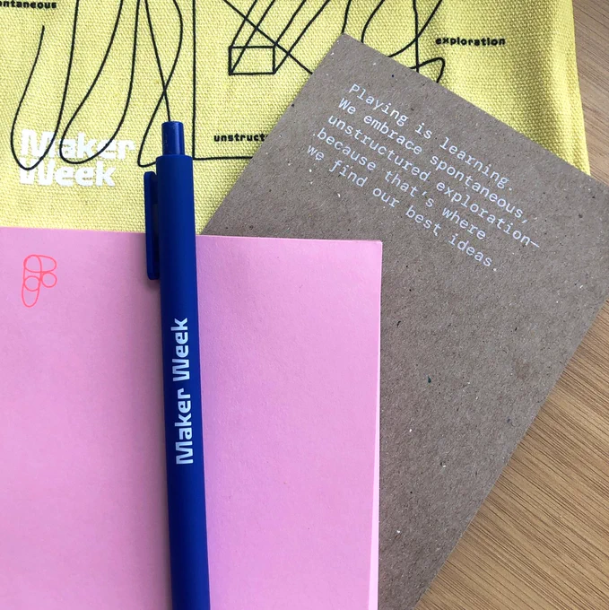 blue pen that says Maker Week sitting against pink notebook with darker pink figma logo. Cardboard says Playing is learning. We embrace the spontaneous unstructured exploration- because that's where find our best ideas.