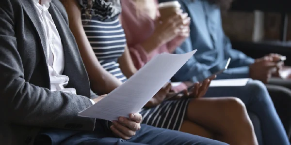 A row of people sits, as if awaiting a job interview. They are professionally dressed, holding papers and electronic tablets.