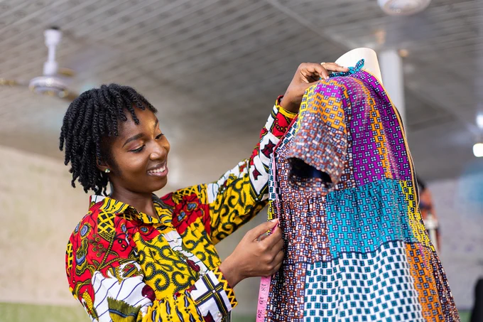 A Black African businesswoman uses a tailor's tape measure to measure a dress on a mannequin.