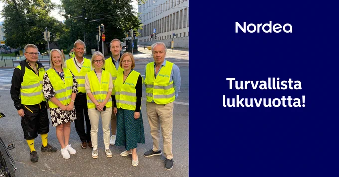 Seitsemän henkilöä seisoo jalkakäytävällä kirkkaisiin huomioliiveihin pukeutuneina ja katsoo hymyillen kameraan. Teksti "turvallista lukuvuotta!". 