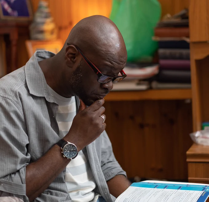 Roger is a stroke survivor wearing glasses and reading from something in his lap in his home. He has one hand on his chin. 