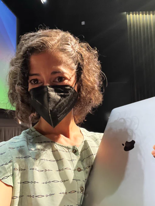 Selfie. Woman with brown curly hair and brown eyes in light teal shirt, wearing black mask and holding a silver Apple laptop. In the background is a huge screen where the presentations will happen. 