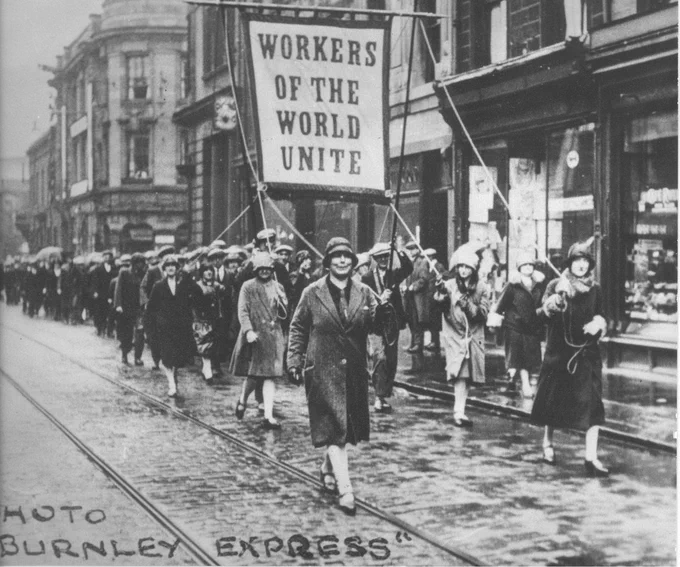 Black and white photo of protesters walking down a street holding a banner that reads 'Workers of the world unite'
