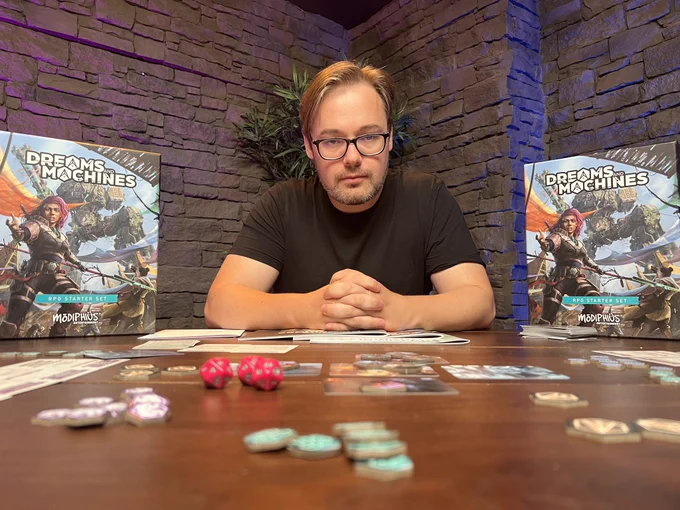A man (mark Hulmes from HighRollers) sat between two box’s with colourful artwork of a ancient mech and pink-haired warrior figure, with the title Dreams & Machines. On the table in front of them is a collection of dice, cards and tokens.
