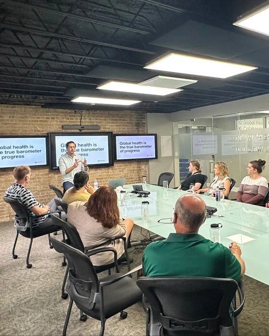 A group of teachers sit at a large table. One person stands at the front the table, with a presentation behind them that reads, “Global health is the true barometer of progress.”