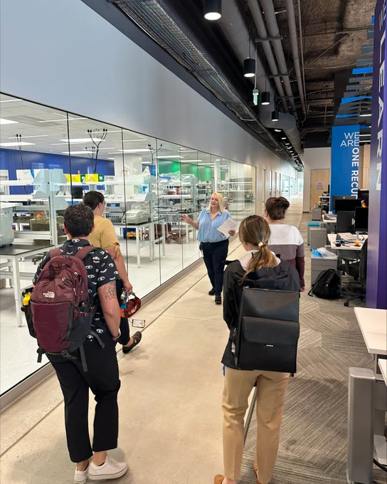Four teachers are given a tour of a lab. A lady at the front of the group is turned around speaking to them. The group is standing in a hall way where a lab can be seen to their left through a glass wall. 
