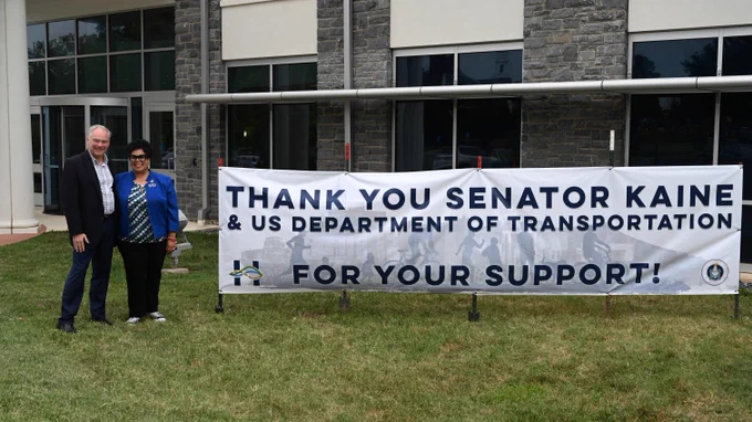 Senator Kaine and Harrisonburg Mayor Reed standing next to a sign that says "THANK YOU SENATOR KAINE & US DEPARTMENT OF TRANSPORTATION FOR YOUR SUPPORT!"
