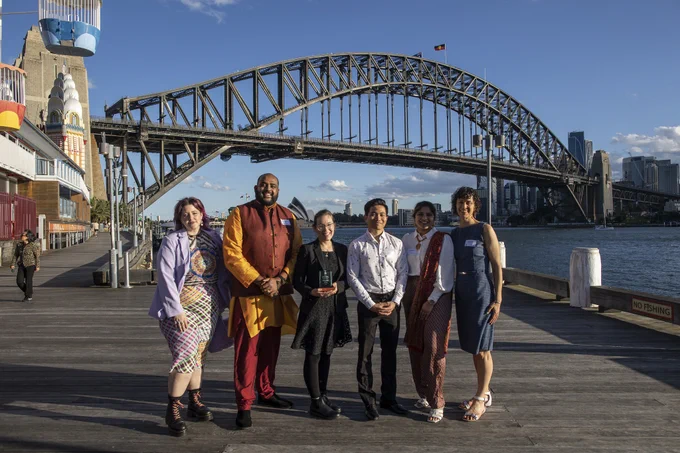 People in colourful clothing in front of the sydney harbour bridge on a wooden wharf