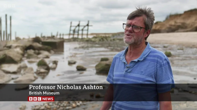 A man in a stripey blue polo shirt standing on a beach. In the background to the right of the picture are cliffs. To the left of the picture are boulders and broken wooden sea defences. A banner across the picture reads "Professor Nicholas Ashton. British Museum. BBC News."