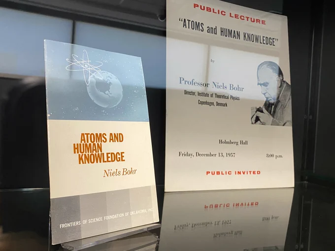 Photo of booklet and poster in display case, both relating to Niels Bohr's 1957 public lecture "Atoms and Human Knowledge"  delivered at the University of Oklahoma.