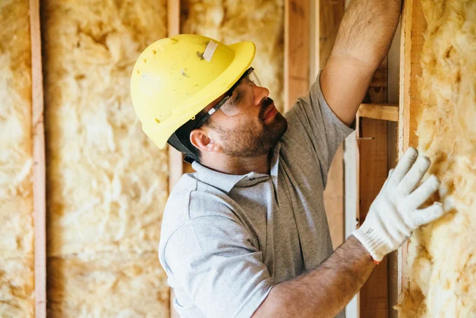 Builder in a yellow hard hat, installing insulation in a wall.