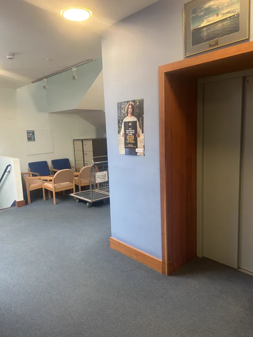 A storage/communal room. There is a Life Changing Poster on the wall next to a lift. The poster reads 'My neighbour did unpaid work and now we share pride in our parks'