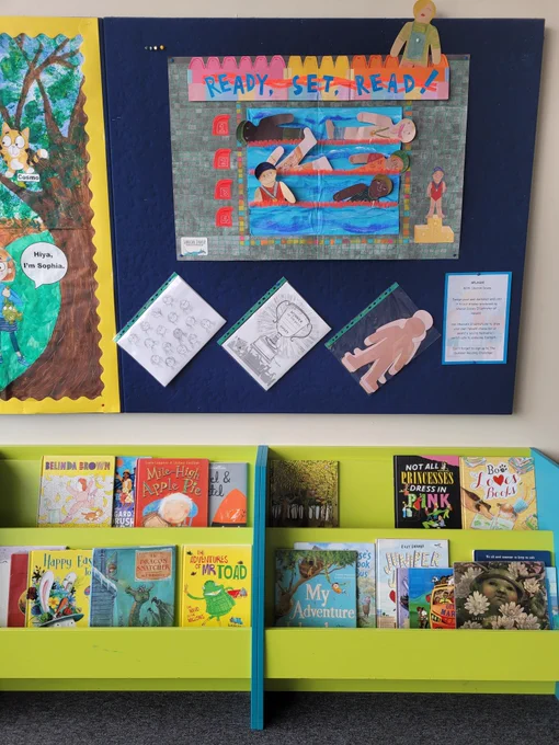 Foreground are some books displayed in oldham children's library on green and blue low shelves. Behind is a display that reads 'Ready, Set, Read!  With a picture of a swimming pool, full of paper cut out swimmers designed by children. 