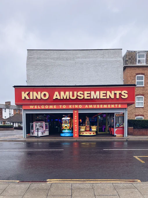 Photograph of the front of a building across a wet road, with 'KINO AMUSEMENTS, WELCOME TO KINO AMUSEMENTS, WIN, WIN, WIN, WIN' in yellow text on red backgrounds, with arcade games visible in the entrance. There's a grey sky above.
