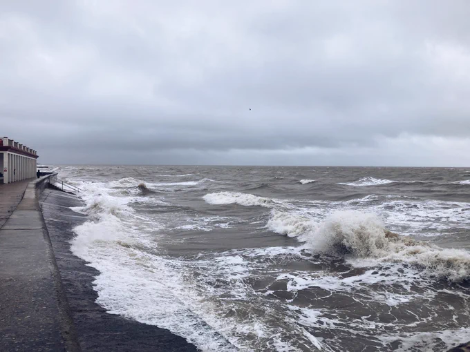 Photograph of the seafront at Walton on the Naze, with concrete walkway to the left, large waves to the right and grey sky above.