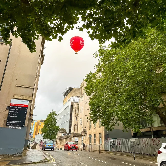 A red hot air balloon is in the sky above the front entrance of Bristol Royal Infirmary and the Bristol Royal Hospital for Children. Green trees frame the image.