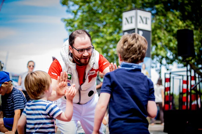 A man wearing glasses, a red top and a white gilet and trousers is leaning down to high five two children. 