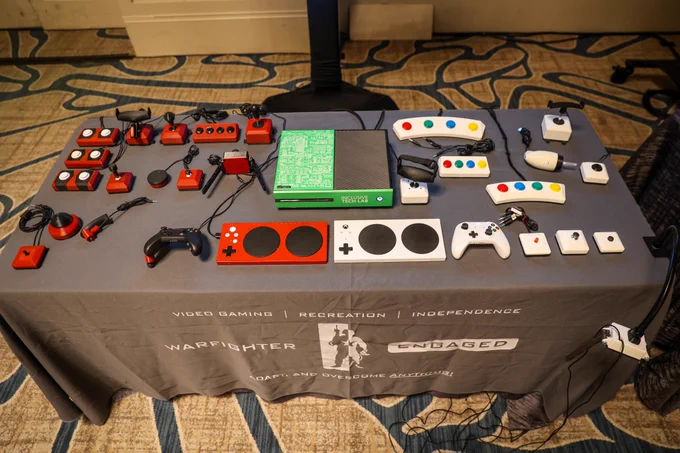 This picture shows a table with an xbox surrounded by multiple buttons and switches that are made by warfighter engaged. The buttons and switches are red and white. The table is dressed with a Warfighter Engaged covering.