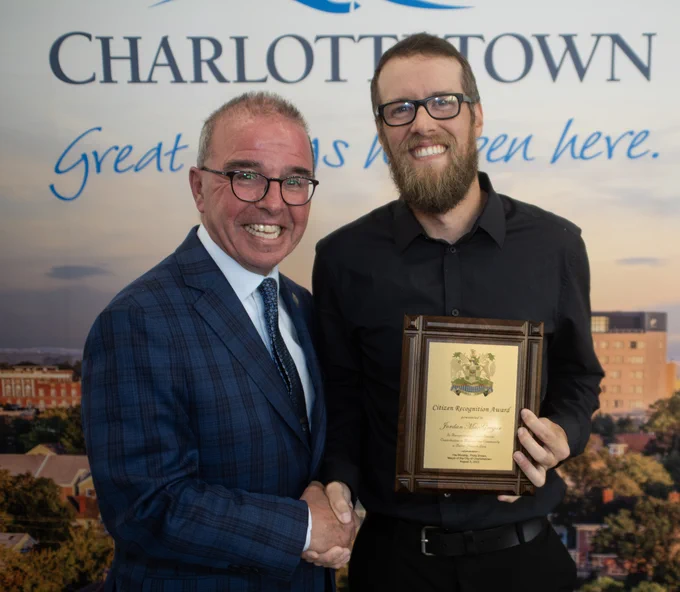 Mayor Philip Brown shakes hands with Jordan MacGregor who is holding his Citizen’s Recognition Award. They are both smiling and posing for the camera in front of a Charlottetown, Great Things Happen Here banner.