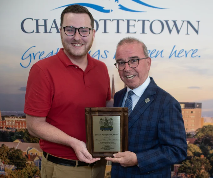 Mayor Philip Brown and a representative of the Kiwanis Club of Charlottetown hold a Citizen’s Recognition Award. They are both smiling and posing for the camera in front of a Charlottetown, Great Things Happen Here banner.