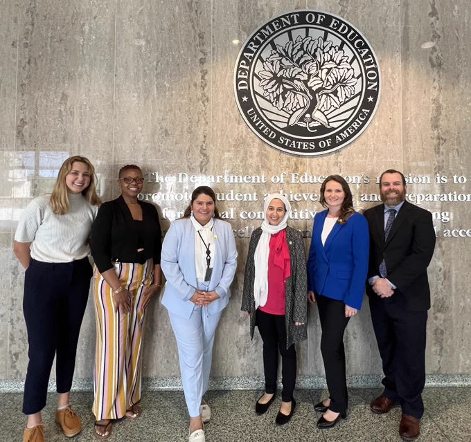A picture of the Summer 2023 team of the Center for Faith Based and Neighborhood Partnerships at the US Department of Education, smiling in front of the Department of Education seal. Left to right are Sylvia Cassidy, Angel Rush, Jasmin Chavez, Maggie Siddiqi, Katy Joseph, and Rob Taber