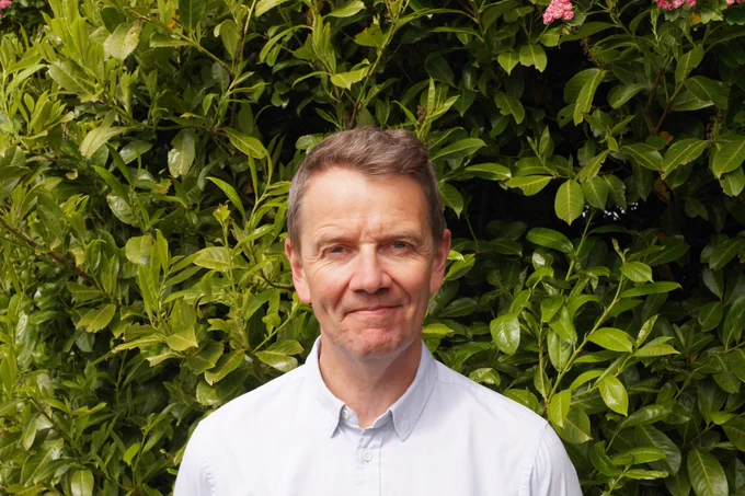 Stephen Lock in a blue smart casual shirt stood in front of a bush full of shiny green leaves.