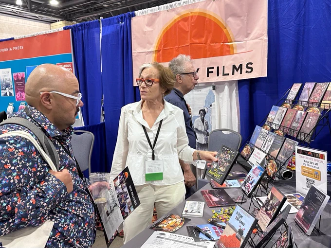 In center, a white woman with a conference name tag is engagingly speaking  about a DVD to a man in a lively flowered shirt, while another man looks at a rack of DVDS. 
