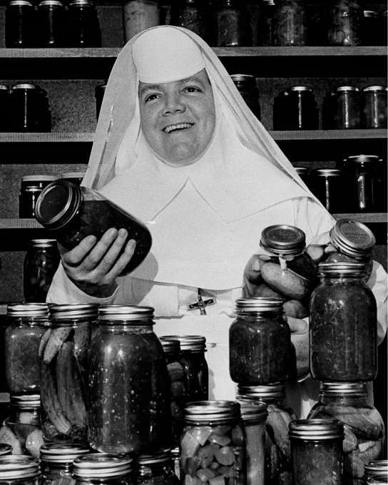 Sister Mary Patricia Doyle, a Religious Sister of the Sisters of Mercy, is holding three glass canning jars of picked cucumbers with many more jars of canned food stacked in front of her and on shelves behind her. The picture is black and white. Sister is wearing a white habit with white veil and crucifix around her neck. She is looking up and smiling at the camera.