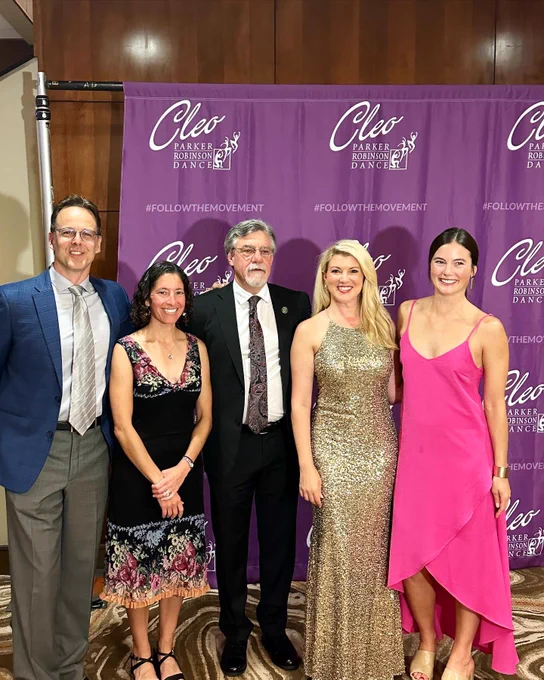 Five people dressed in suits and dresses pose in a line together in front of a purple Cleo Dance step and repeat backdrop. From left to right it is Noah Racey, Emily Morgan, Dean Ben Withers, Amy Parsons and 
Grace Gallagher.