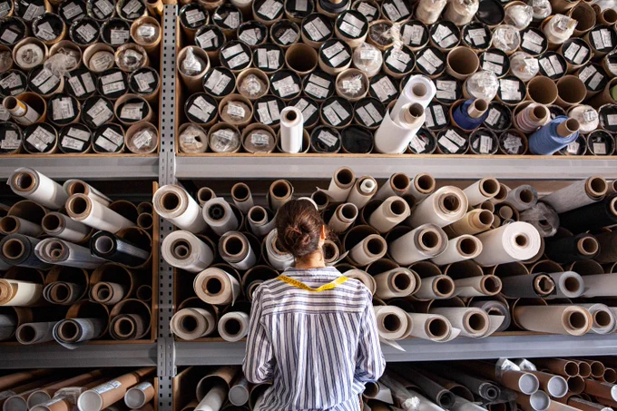 A person with a measuring tape draped around their shoulders looks at shelves filled with rolls of fabrics