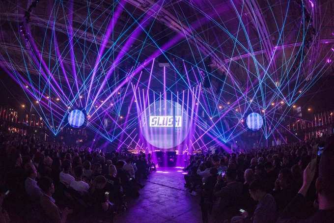Dark room with people sat looking at a stage with a spotlight that says 'Slush'. There are lots of blue and purple laser lights crossing over the stage.