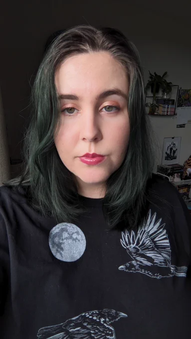 A white woman is failing to smize into the camera. She has a shirt with crows on it, burgundy lipstick, and dark green hair. 
