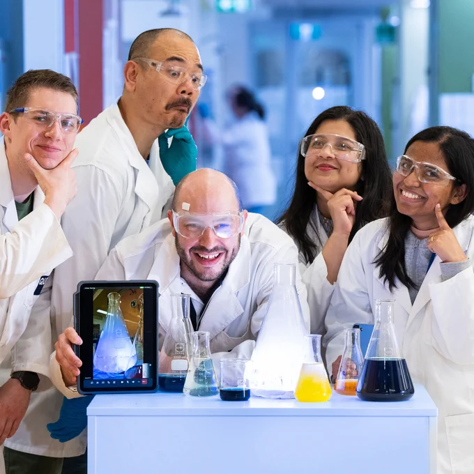 3 x male and 2 x female science teachers doing fun poses in a group in a lab with lab equipment in front of them, wearing safety glasses
