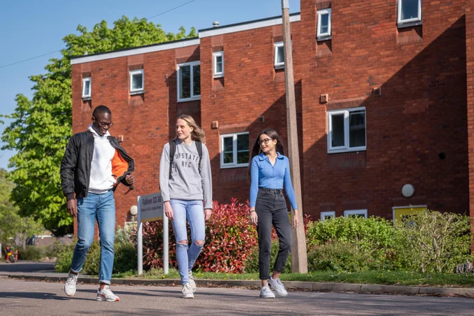 Three students walking on campus in front of an accommodation building. They also have flowers and trees surrounding them.