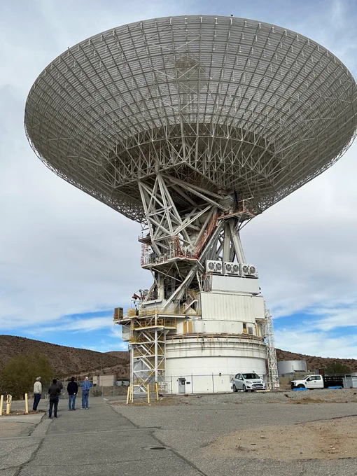 This is the 70 meter antenna at Goldstone, California. The dish is enormous, taking up most of the image. The dish, which is white with lines running through it is turned up to the sky. It has a white base that attaches it to the ground. Four people stand to the left giving a sense of scale.  Behind it are low mountains and a mostly cloudy sky.