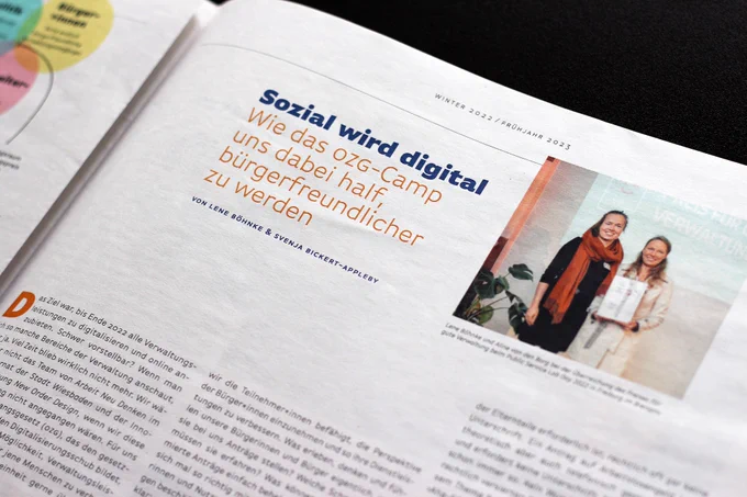 Detail einer Zeitungsseite, die ein Bild mit 2 Frauen auf einer Bühne zeigt, neben ihnen die Überschrift über einem längeren Text: ›Sozial wird digital – Wie das OZG-Camp uns dabei half, bürgerfreundlicher zu werden‹