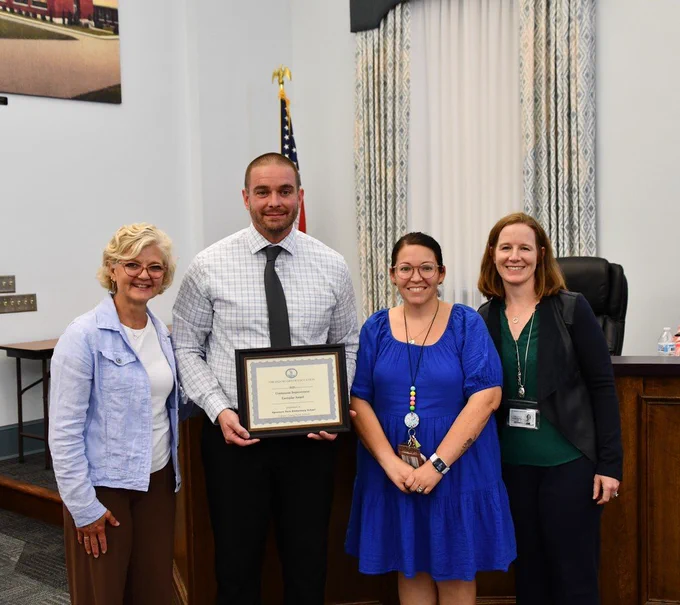four people smiling in board room, one holding an award certificate