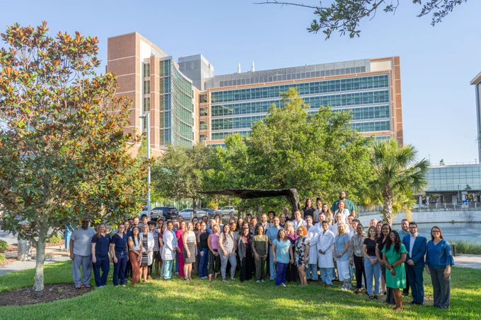 Dozens of University of Florida and UF Health employees in the abdomnial transplant teams stand to the right of a tall magnolia tree outside the UF Health hospital in Gainesville, Florida, for a group photo.