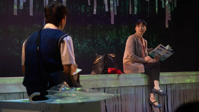 A young woman sits on a wall, reading a book. She has her hair tied back, wears a beige blazer, smart black trousers and heels, and looks over to a young boy sat in the foreground.