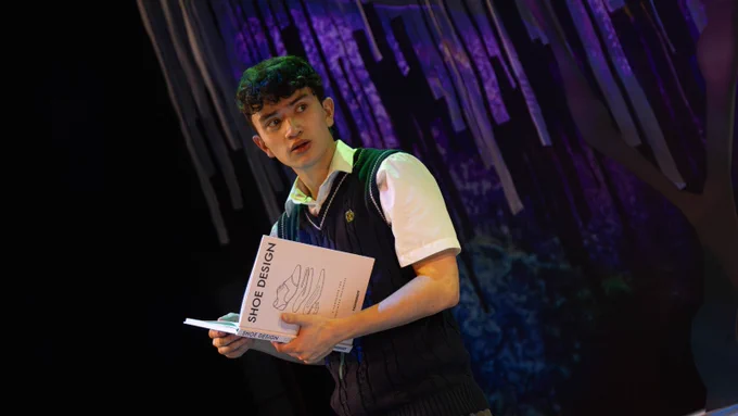 A young man, wearing a school sweater vest over a white shirt. He holds a book titled 'shoe design'. Behing him, long branches of a willow tree hang behind him, illuminated in purple lighting. 