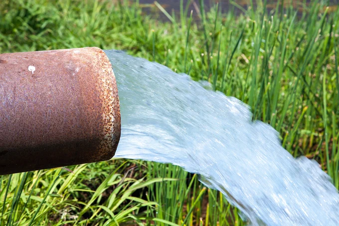 A large rust colored pipe has groundwater flowing out of into a green rice field.